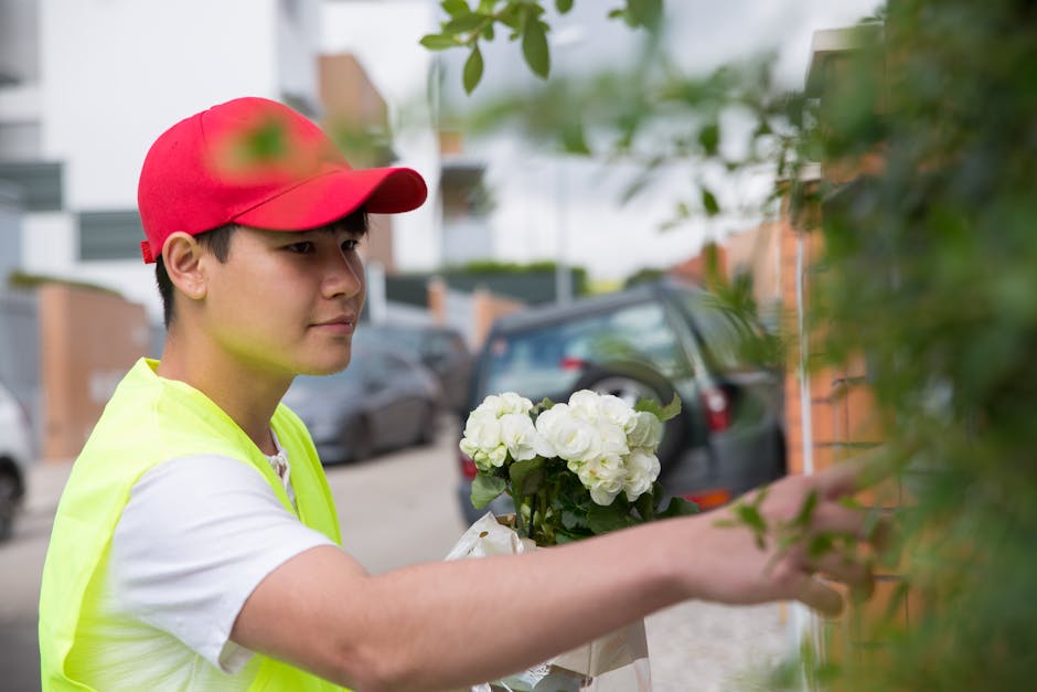A young male florist wearing a red cap and a neon yellow safety vest arranges a bouquet of white roses and green foliage against a brick wall, with parked cars and residential buildings visible in the
