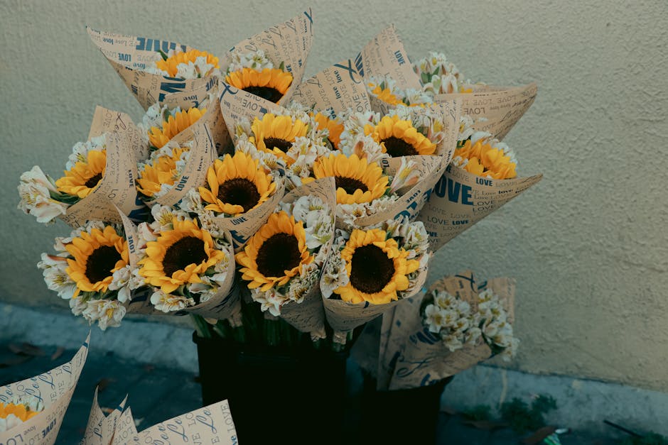 A collection of fresh sunflower bouquets with bright yellow petals and dark centers, wrapped in paper printed with blue and beige text including words like 'LOVE,' arranged in a black container agains