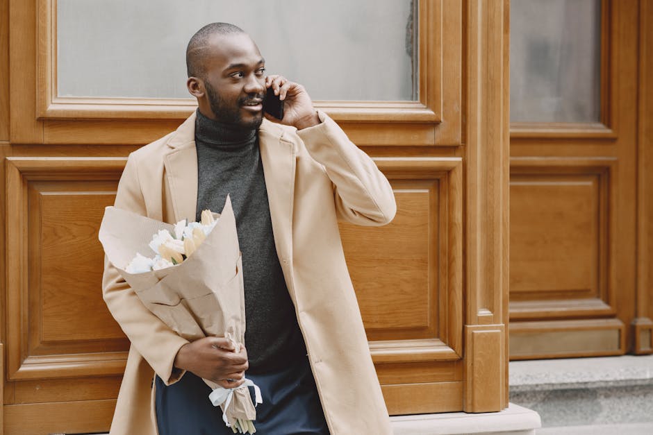 A man with dark skin, a neatly trimmed beard, and a shaved head, wearing a beige overcoat and a dark grey turtleneck, is standing outdoors against a backdrop of wooden paneling. He is holding a bouque