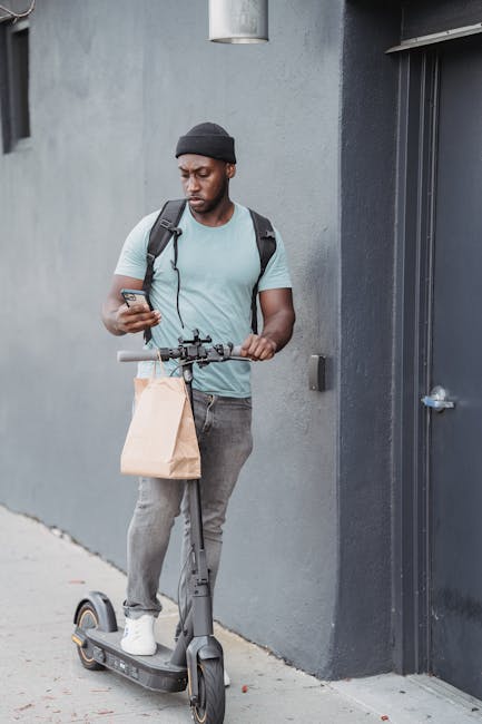 A young man with dark skin and a beard stands beside a gray wall, holding a smartphone in one hand and gripping the handlebar of an electric scooter with the other. He is dressed casually in a light b