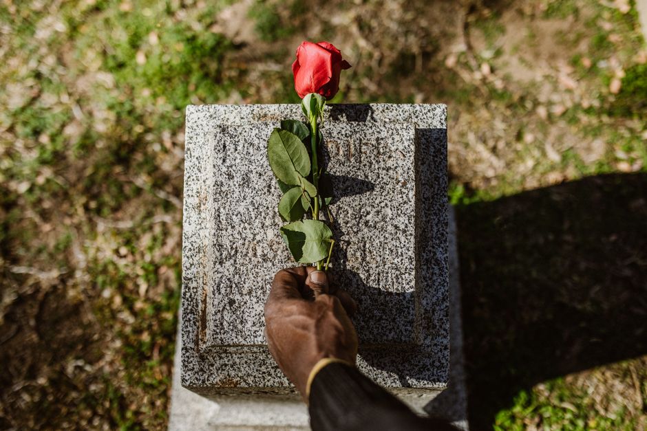 A close-up image of a person's hand holding a single red rose with green leaves, positioned above a granite memorial stone. The stone is textured and gray, set outdoors with sunlight casting shadows,