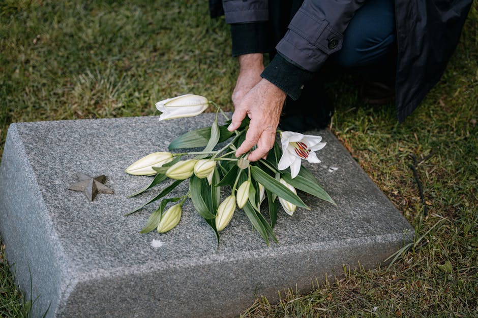 A person placing a bouquet of fresh white lilies and unopened lily buds on a granite memorial plaque with a small engraved star, outdoors on a grassy surface. The bouquet features large white blooms w
