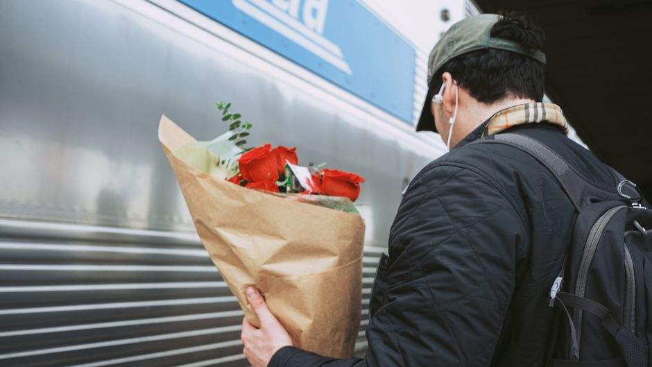 A person dressed in a dark jacket with a plaid scarf, gray cap, and earbuds, holding a bouquet of fresh red roses wrapped in brown kraft paper, standing outdoors near a metallic delivery vehicle with