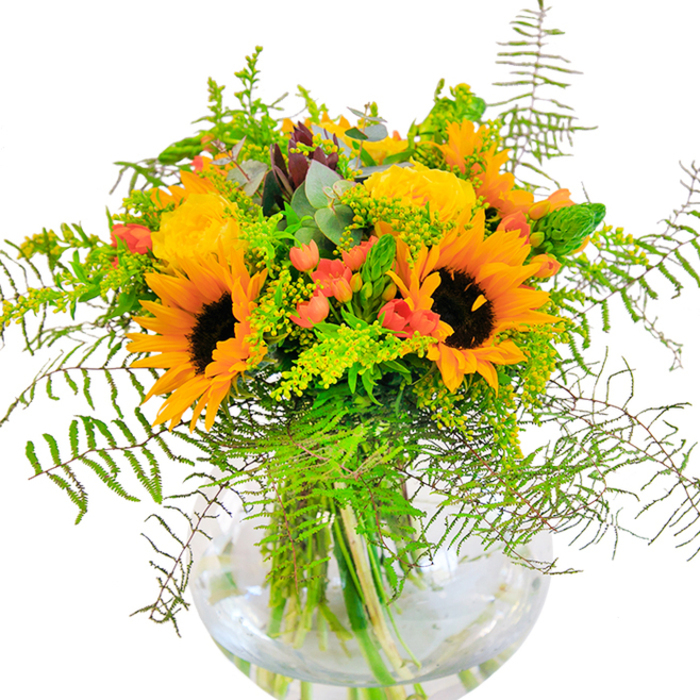 The impression left by this radiant bouquet is one of joyous abundance, reminiscent of a walk through the wildflower-rich stretches on the fringes of Well Street Common in Homerton. Majestic sunflowers, their wide faces brimming with energy and bordered by luminous yellow-orange petals, command attention at the heart of the arrangement. Intermingled are sumptuous golden garden roses, their tightly wound petals creating plush, intricate spirals reminiscent of vintage romance. Glossy red Hypericum berries peek from within the foliage, their bold colour and rounded form popping amidst the softer textures, much like the vibrant community spirit along Homerton's lively streets. A profusion of greenery, led by airy, featherlike fern and sprays of tiny yellow solidago, spill from the arrangement's edges, bestowing a sense of organic motion and freshness. Delicate, powdery blue-green eucalyptus leaves infuse calming undertones, suggesting a crisp, invigorating scent. Captured in a transparent, fishbowl-like vase, each stem is visible, echoing the sincerity and skill of local Homerton artisans. Light reflects off the water, highlighting the contrasting shapes and colours within. This bouquet is the perfect emblem for all manner of celebrations in E9-from birthdays enjoyed at home to gatherings near the artistic centricity of Hackney Wick-and brings a zestful, nature-inspired beauty that lifts any space. The bouquet's harmonious medley of colours and textures tells a story of late-summer festivity and heartfelt connection, right from the heart of the community.